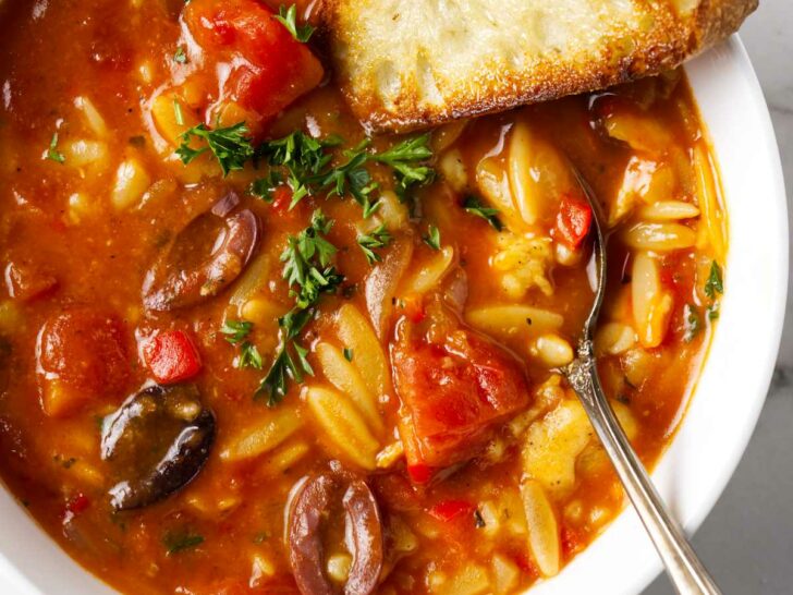 A spoon in a bowl of chicken cacciatore orzo soup along with a slice of French bread.