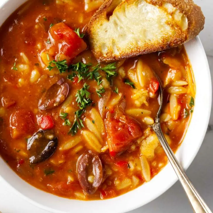 A spoon in a bowl of chicken cacciatore orzo soup along with a slice of French bread.