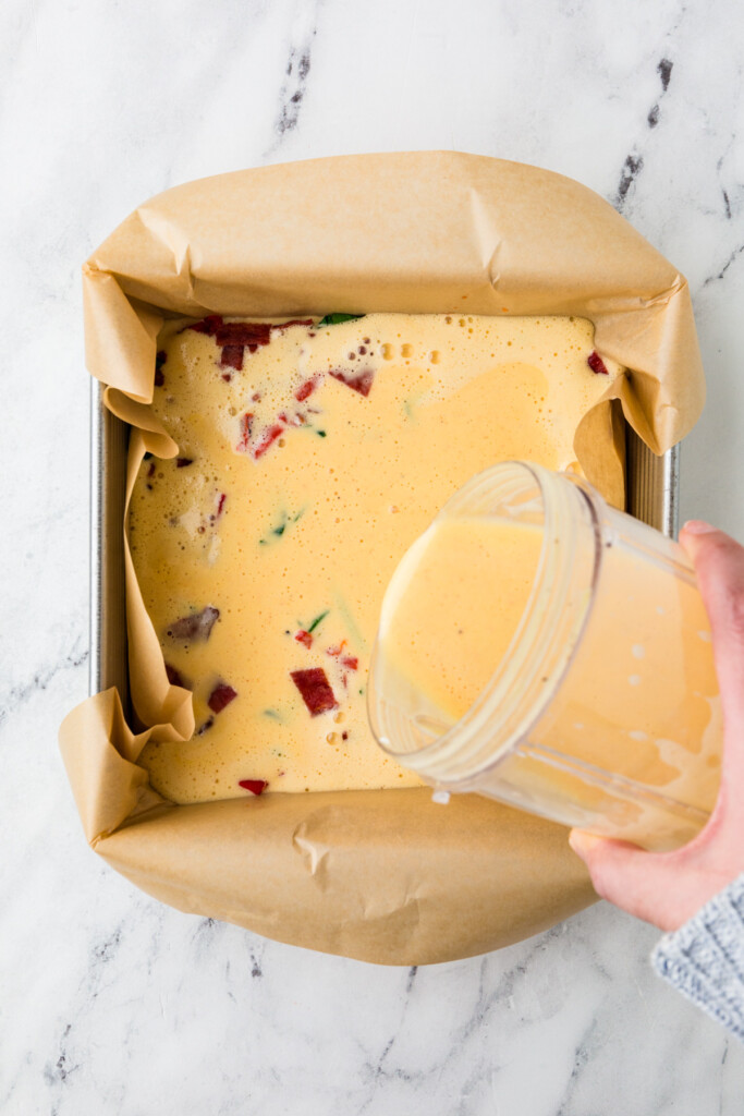 Pouring egg cottage cheese casserole mixture into a baking dish with veggies.