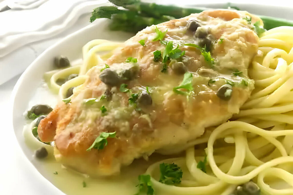 A chicken breast with lemon caper sauce on a bed of pasta.