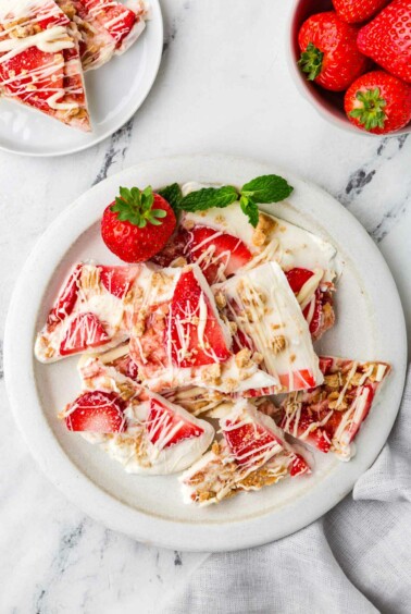 A plate with filled with thin wedges of frozen yogurt bark next to some fresh strawberries.