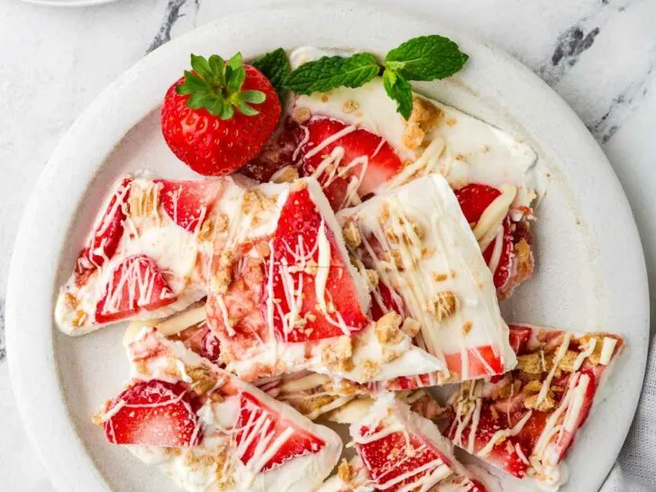 A plate with filled with thin wedges of frozen yogurt bark next to some fresh strawberries.