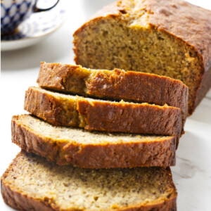 A loaf of banana bread with four thick slices in front of a cup of coffee.