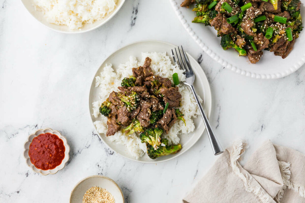 A beef stir fry with broccoli on a plate next to rice and hot sauce.