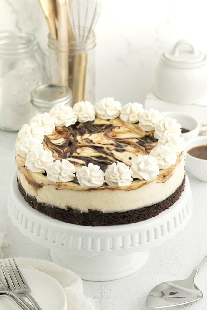 A brownie bottom cheesecake with swirls of chocolate and caramel sitting on a cake plate next to a spatula.