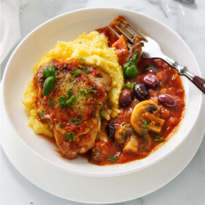 A white dish with Italian chicken cacciatore with mushrooms and olives over a bed of polenta.