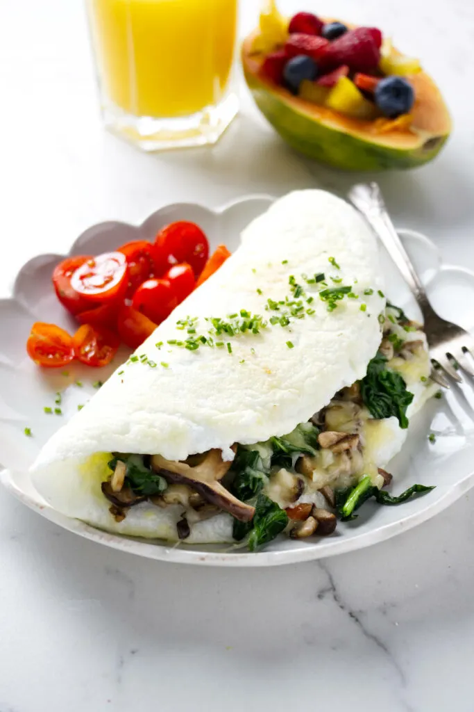 A fluffy egg white omelet filled with mushrooms and spinach with fruit and orange juice in the background.