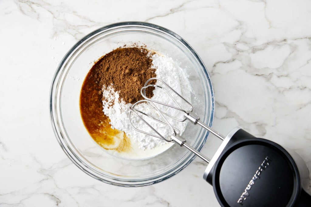 Sugar, cocoa, vanilla and cream in a bowl next to a mixer.