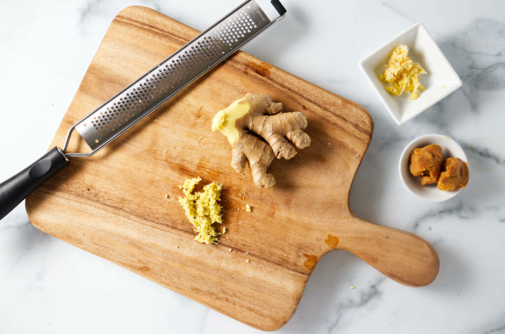 Cutting board with grated ginger, ginger root and micro-grater. Small dish of grated garlic and a dish with miso.