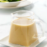 Close up photo of a pitcher of Miso-Ginger Dressing sitting on a small plate. A dish of salad in the background.