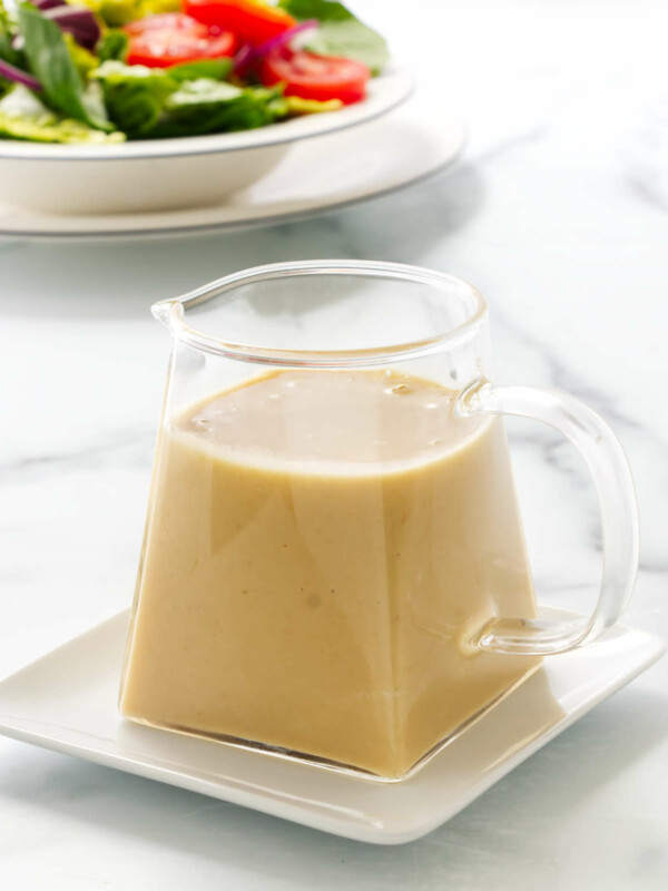 Close up photo of a pitcher of Miso-Ginger Dressing sitting on a small plate. A dish of salad in the background.