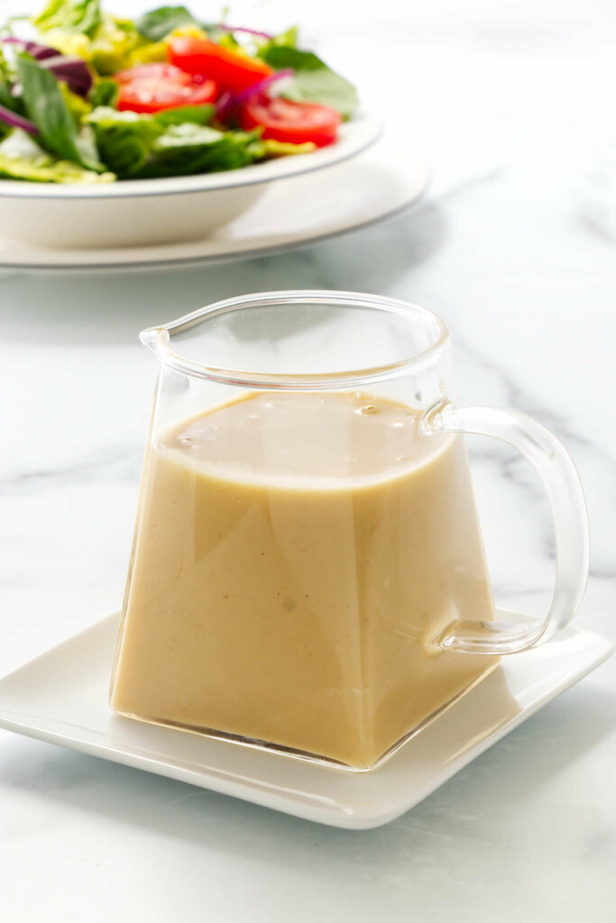 Close up photo of a pitcher of Miso-Ginger Dressing sitting on a small plate. A dish of salad in the background.