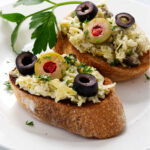 Two artichoke bruschetta on a plate topped with olives.