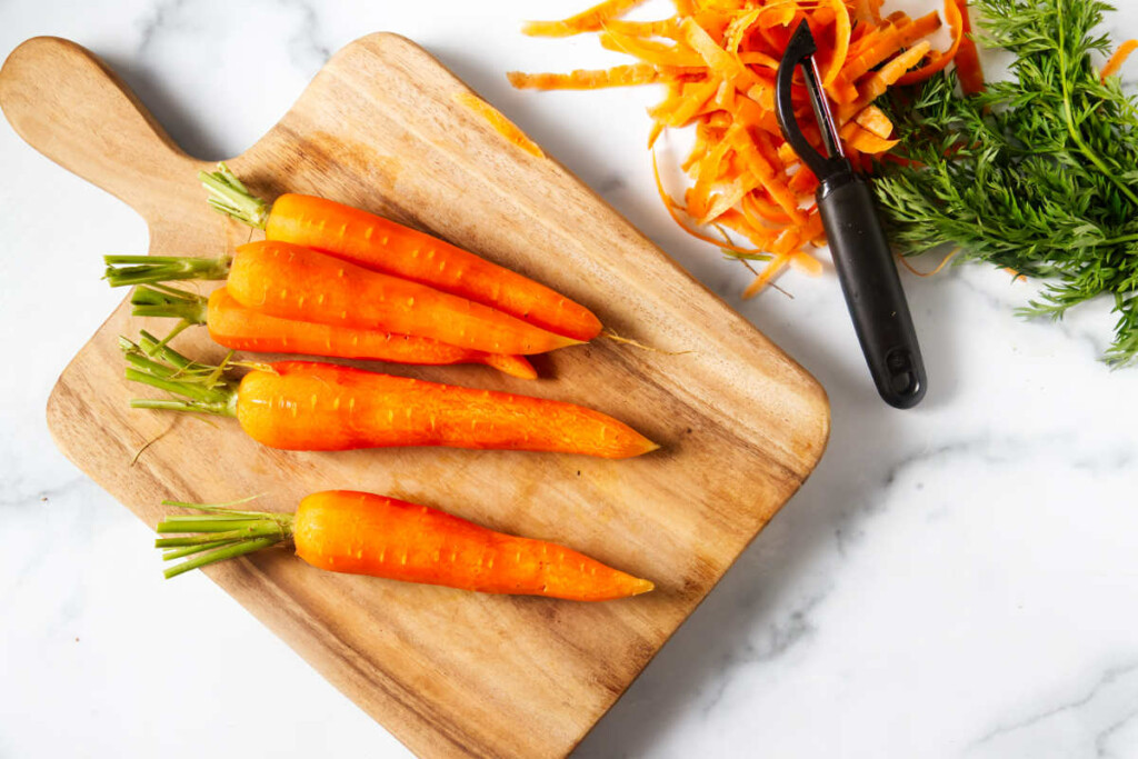 Several peeled carrots next to a vegetable peeler and carrot tops.