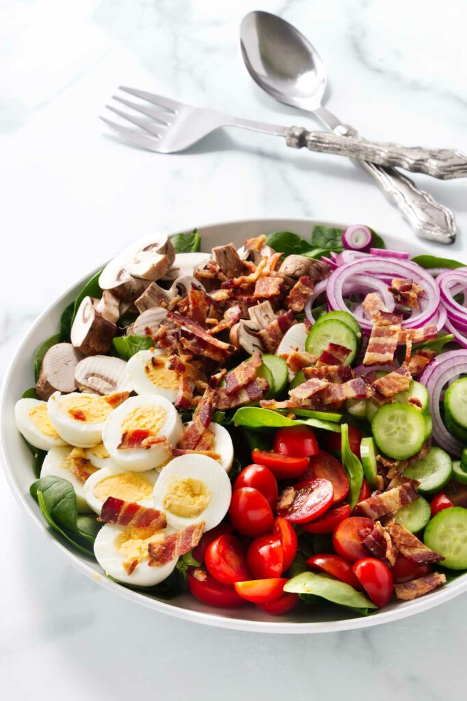 A bacon and tomato spinach salad next to a large spoon and fork.