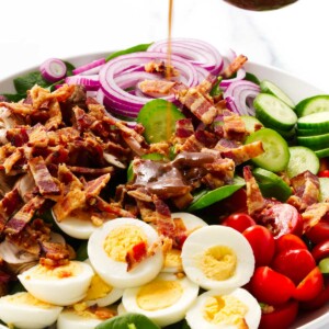 Pouring balsamic vinaigrette over a spinach bacon salad with tomatoes and eggs.