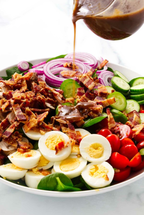 Pouring balsamic vinaigrette over a spinach bacon salad with tomatoes and eggs.