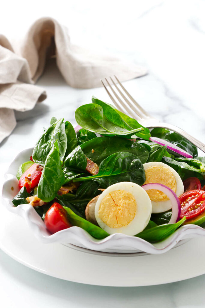 A salad plate with spinach, hard boiled eggs, bacon, and veggies.