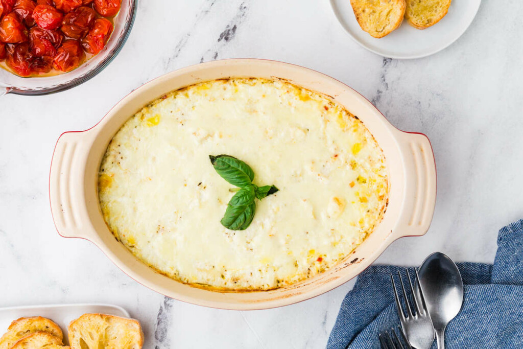 A warm ricotta spread recipe in a baking dish next to roasted tomatoes.