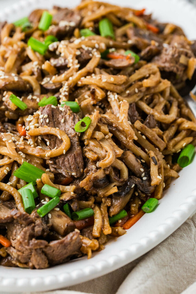 Steak udon with noodles on a dinner plate topped with sesame seeds and scallions.