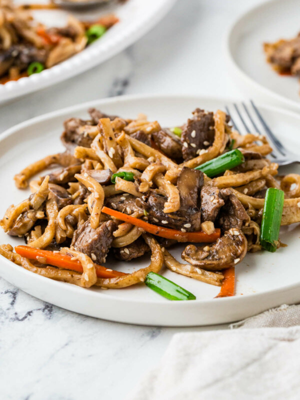 Beef udon noodles on a white plate next to a serving platter.