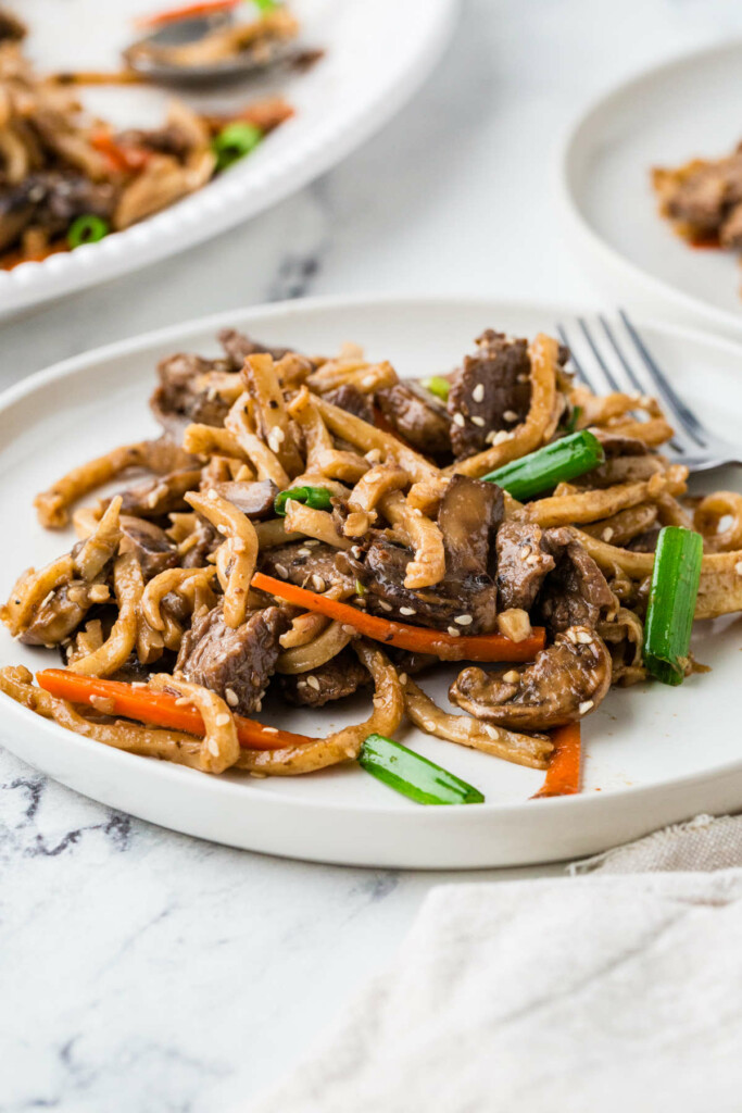 Beef udon noodles on a white plate next to a serving platter.