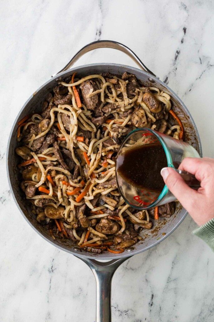 Adding stir fry sauce to udon beef noodles in a skillet.