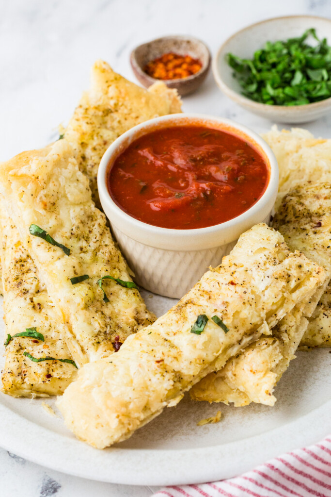 Homemade cheese breadsticks on a plate with tomato sauce; herbs in the background.