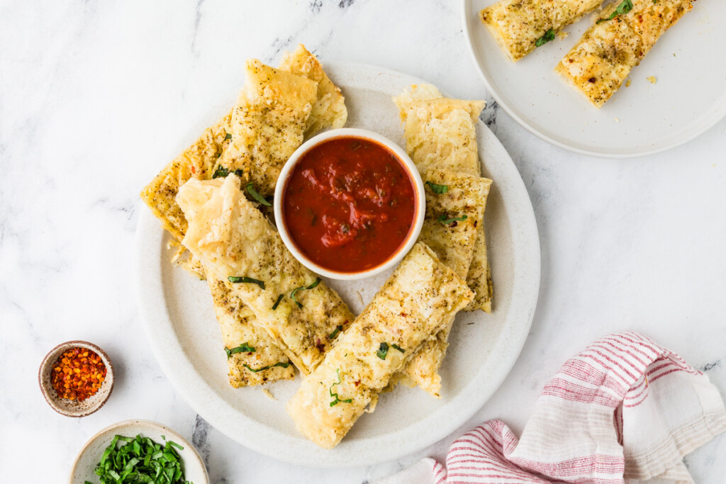 A plate filled with pizza crust breadsticks and a bowl of marinara sauce.