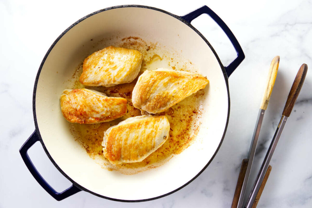 Four browned chicken breasts cooking in a white pot with blue handles.