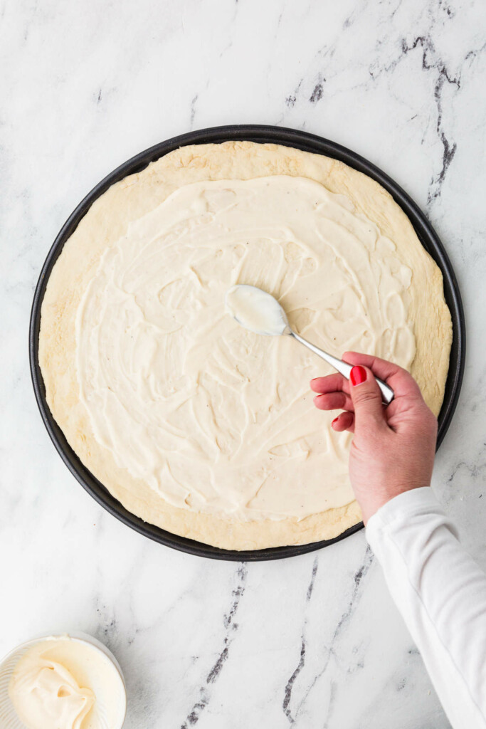 A hand with red nails spreading white sauce on pizza dough.