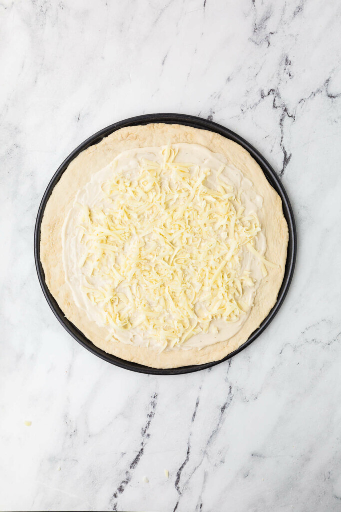 Pizza dough with white sauce and cheese on a black tray.