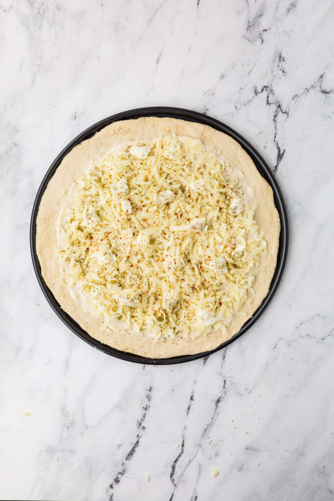 Unbaked pizza crust with cheese and herbs on black tray.