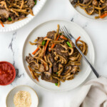 Beef udon noodles on a dinner plate next to chili garlic sauce.