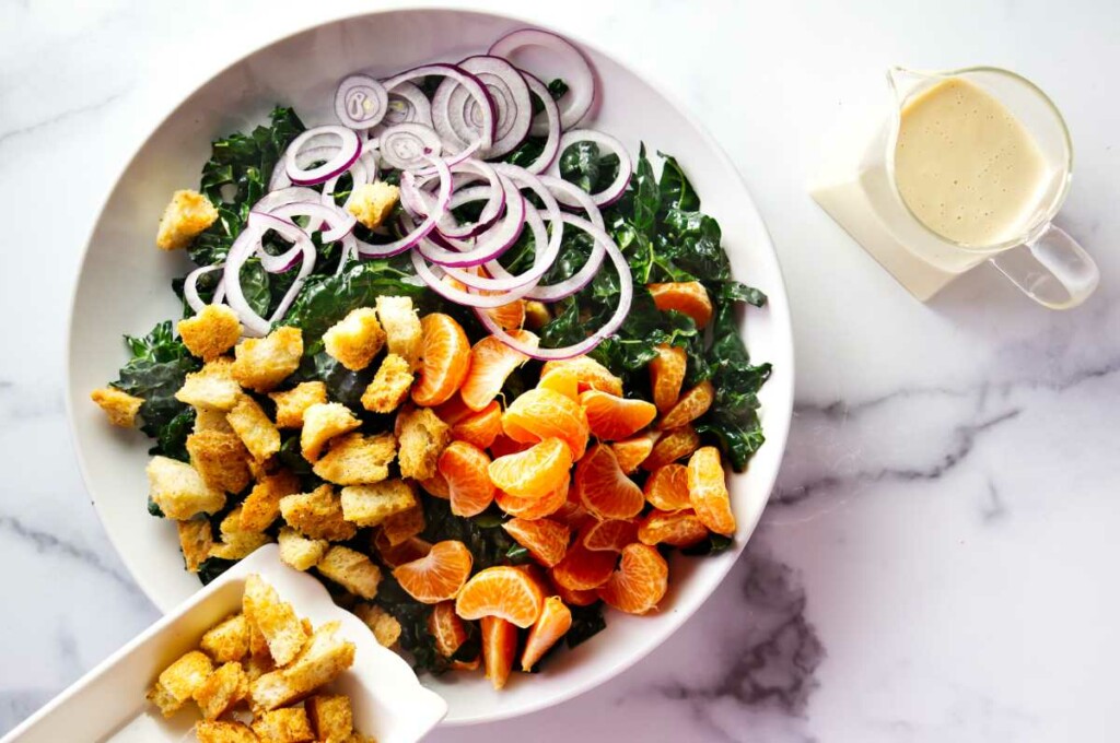 Adding mandarin orange slices, croutons, and onions to a bowl with kale.