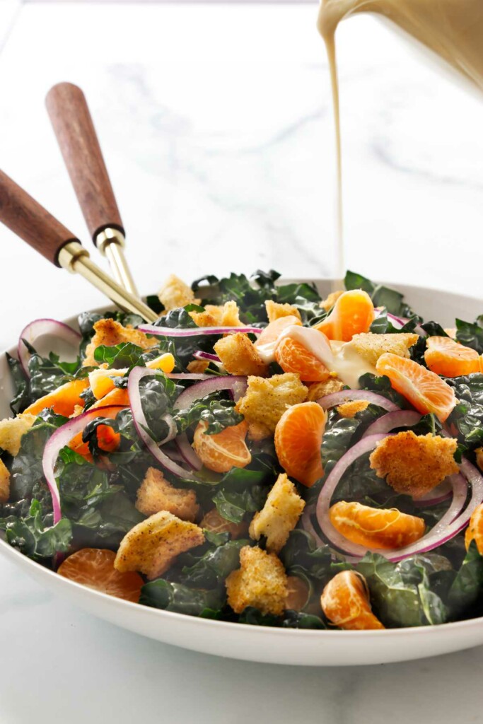 Pouring miso ginger dressing over a kale salad with oranges.