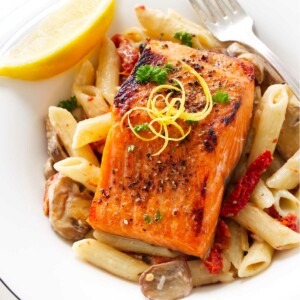 A serving of broiled salmon on a bed of penne pasta, garnished with lemon zest, parsley and a lemon wedge.