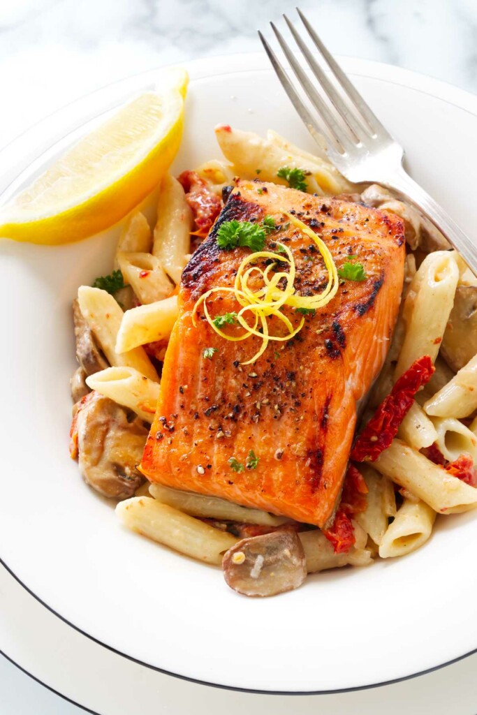 A serving of broiled salmon in a dish of pasta with mushrooms, sun-dried tomatoes and lemon cream.sauce.