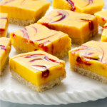 Close-up of several square mango bars with raspberry swirls on a white serving plate. Each dessert features a vibrant yellow mango curd layer intertwined with red raspberry swirls.