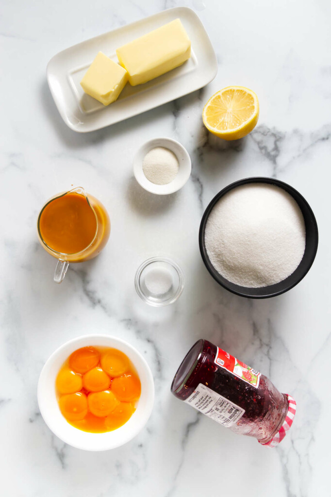 Ingredients on a marble surface: butter on a dish, a halved lemon, a small bowl of white powder, a large bowl of sugar, a bowl with egg yolks, a pitcher of orange liquid, salt in a small dish, and a jar of red jam.