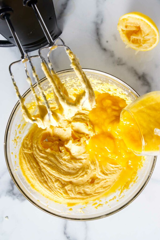 Mixing bowl with creamy batter, hand mixer resting, mango curd being poured in.