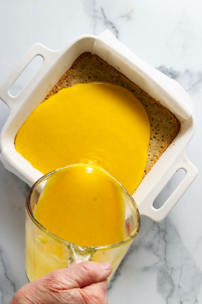 Hand pouring mango curd batter onto a shortbread crust in a baking dish