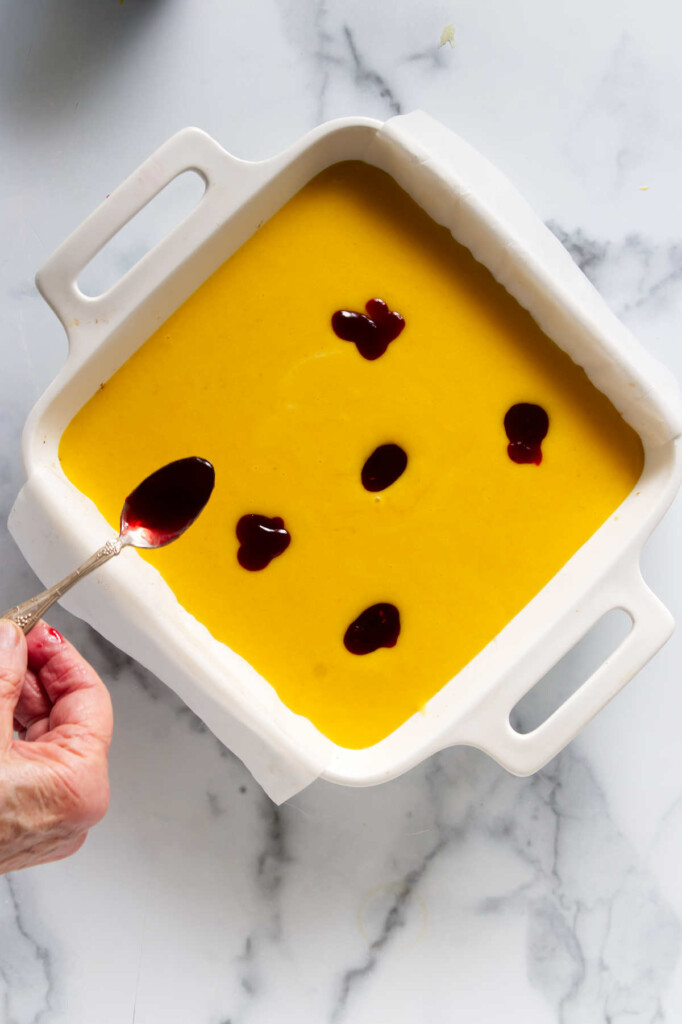 Drizzling raspberry syrup over mango curd batter in a dish preparing mango bars.