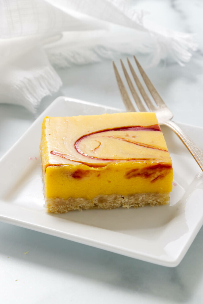 A square slice of a mango bar with a raspberry swirl on top is on a white plate.