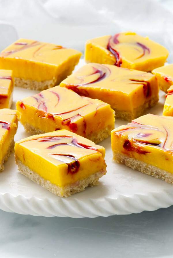 Nine squares of layered mango dessert bars sit on a white plate. The bars have a marbled raspberry top, with a creamy layer over a crumbly base.