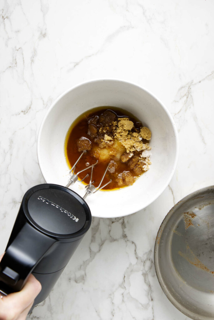 Hand mixer blending brown sugar and wet ingredients in a white bowl on a marble countertop.