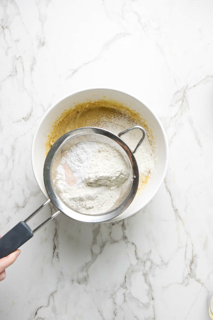 Sifting flour over white bowl with sourdough discard cookie batter.