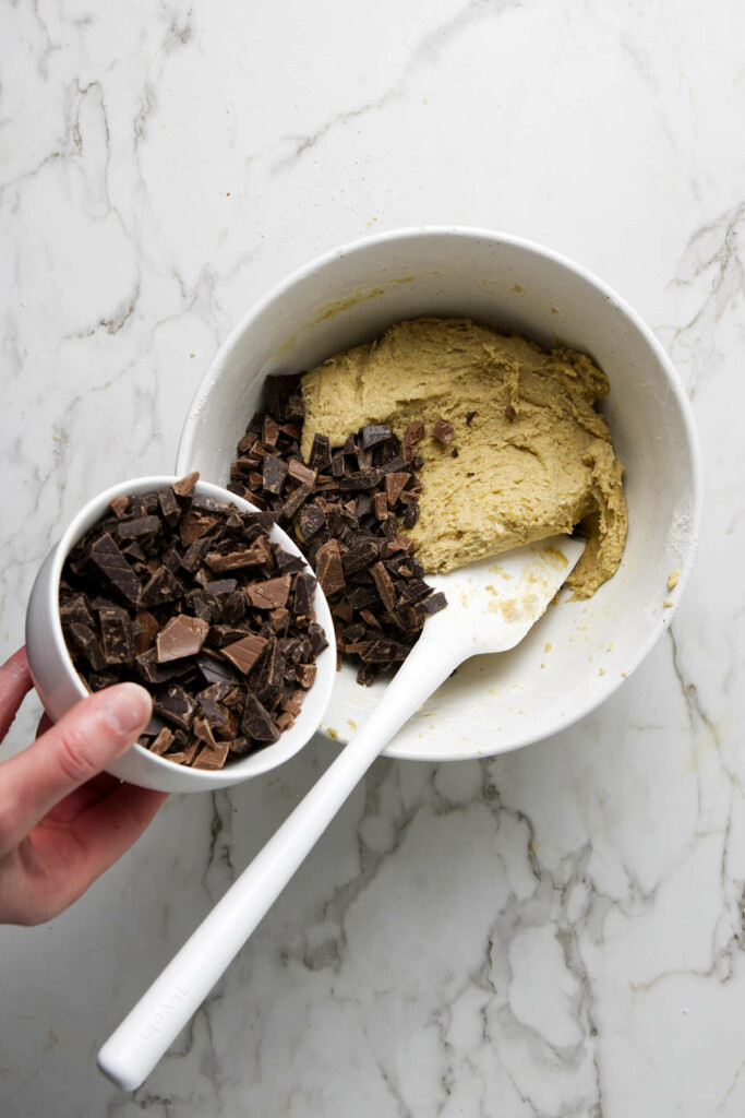 Adding chocolate chunks to sourdough cookie dough in a white bowl.