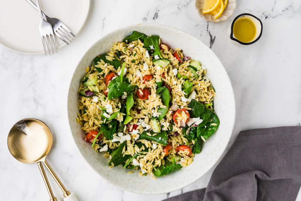 An orzo and feta salad in a serving bowl next to salad utensils.