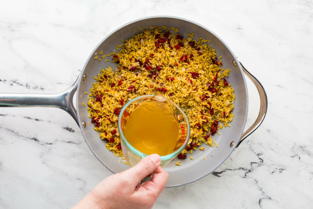 Pouring chicken broth in a skillet with orzo and tomatoes.